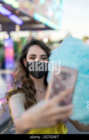 Donna in maschera protettiva e con caramelle di cotone dolce blu che si autosshot sul telefono cellulare mentre si divertono al campo da golf Foto Stock