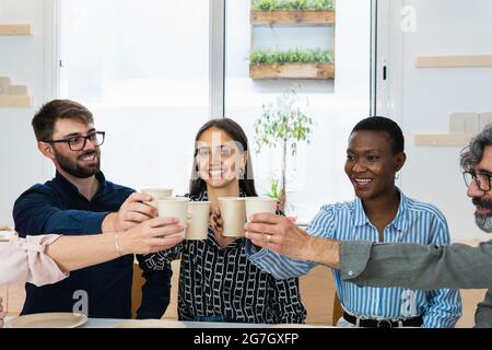 Gruppo di collaboratori multirazziali che si riuniscono a tavola e a tazze di clinking mentre si pranzano insieme in cucina sul posto di lavoro Foto Stock