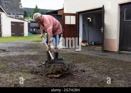 13 luglio 2021, Sassonia, Jöhstadt: Un uomo tira via lo sporco dopo un'inondazione nel distretto di Jöhstadt di Steinbach, nel Erzgebirgskreis. Il giorno dopo l'alluvione, i residenti, il cantiere, i vigili del fuoco e i soccorritori sono impegnati nel lavoro di pulizia. I detriti in entrata continuano a bloccare gli scarichi. Uno dei residenti di Jöhstädt è stato spazzato via dal flash flood, è ancora mancante. Foto: Ronny Küttner/dpa Foto Stock