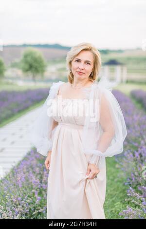 Bella donna bionda matura in elegante abito beige lungo in posa in campo lavanda. Donna di mezza età in abito all'aperto. Foto Stock