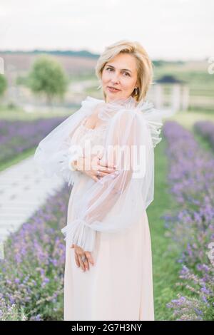 Bella donna bionda matura in elegante abito beige lungo in posa in campo lavanda. Donna di mezza età in abito all'aperto. Foto Stock