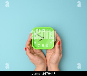 la mano della donna tiene una sveglia verde quadrata, l'ora è dieci - otto alla mattina. Concetto di Early Rise, sfondo blu Foto Stock