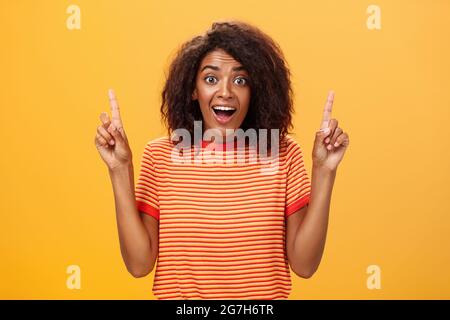 Stupito felice entusiasta donna afroamericana carina con acconciatura riccia in t-shirt a righe che solleva le dita indice puntando su guardare ipnotizzato e. Foto Stock