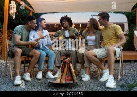 Funky diversi amici che suonano la chitarra e canti vicino al falò, viaggiando in camper, godendo di vacanze estive. I Millennial multirazziali vanno avanti Foto Stock
