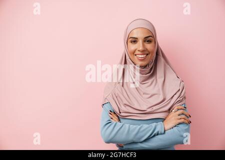 Una donna musulmana sorridente che indossa hijab rosa in piedi con braccia incrociate nello studio rosa Foto Stock