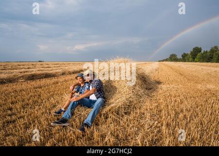 Padre e figlio riposano dopo il raccolto riuscito. Arcobaleno nel cielo dietro di loro. Foto Stock
