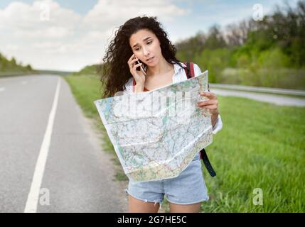 La turista stressata con la mappa in piedi sulla strada, parlando su smartphone, non può prendere l'auto o trovare il suo percorso di viaggio. Giovane donna sentendosi perso whil Foto Stock