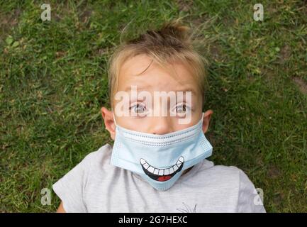 il bambino in una maschera medica blu con un sorriso dipinto su di esso giace sull'erba con ampi occhi aperti. Atteggiamento positivo durante la pandemia di COVID 19. W Foto Stock