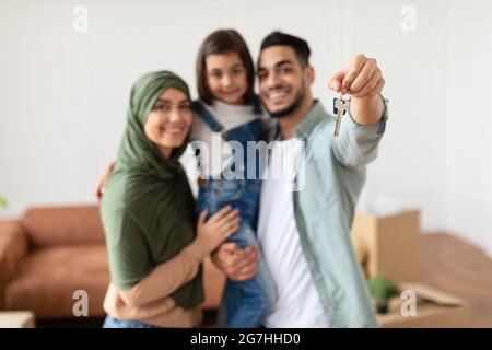 Proprietà della casa. Famiglia musulmana felice che mostra chiave, fuoco selettivo sulla mano. Genitori allegri abbracciando piccolo Kid, persone che si spostano in nuovo appartamento Standin Foto Stock