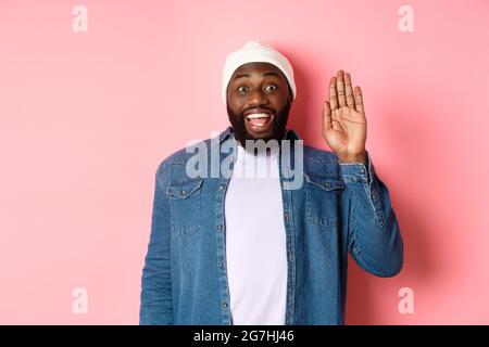 Amico sorridente uomo nero che ciao, con la mano, vi accoglie, in piedi su sfondo rosa Foto Stock