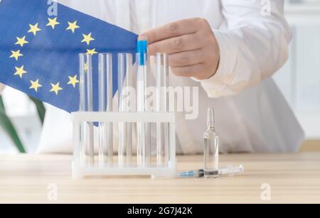 Il medico prende la bandiera dell'Unione europea vicino alla provetta, alla fiala e alla siringa, concetto di vaccinazione globale. Foto Stock