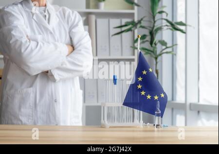 Il medico si trova vicino alla provetta, alla fiala, alla siringa e alla bandiera dell'Unione europea, concetto di vaccinazione globale. Foto Stock