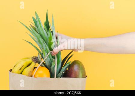 Borsa di carta piena di frutta tropicale con la mano della donna su sfondo giallo. Spazio di copia. Foto Stock