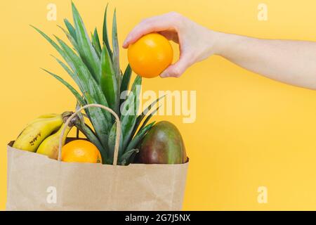 Borsa di carta piena di frutta tropicale con la mano della donna su sfondo giallo. Spazio di copia. Foto Stock