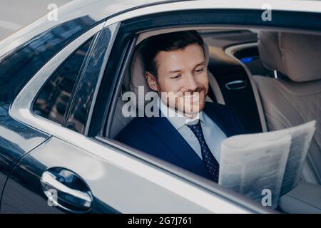 Bel giovane banchiere in tuta elegante formale smoking legge giornale in auto di lusso Foto Stock