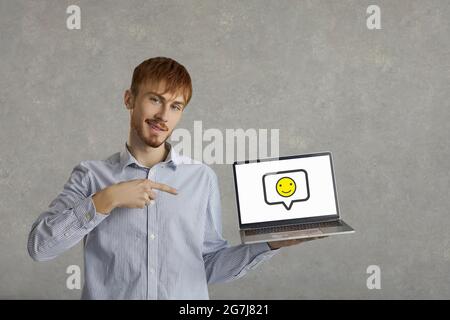 Felice giovane uomo che indica un emoticon sorridente sullo schermo del suo computer portatile Foto Stock