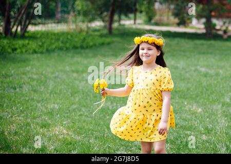 una bella ragazza felice in un vestito che vola nel vento corre sull'erba verde e con i dandelions nelle sue mani e una corona sulla sua testa nel parco su Foto Stock