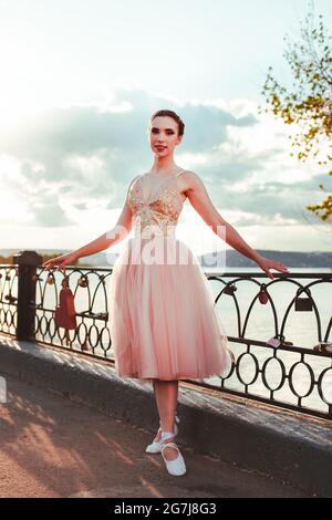 Una giovane ballerina si trova in un abito rosa e cechi con nastri su una ringhiera in ghisa del recinto del fiume in una calda giornata estiva al tramonto Foto Stock