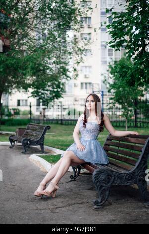 una giovane donna in un abito festoso corto blu corset si siede con grazia su una panchina di legno in un parco cittadino in estate Foto Stock