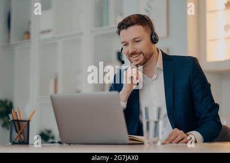 L'operatore maschile professionale lavora in callcenter utilizza cuffie e computer portatile per le conversazioni in videochiamata o evento webcam virtuale indossa abiti formali pose Foto Stock