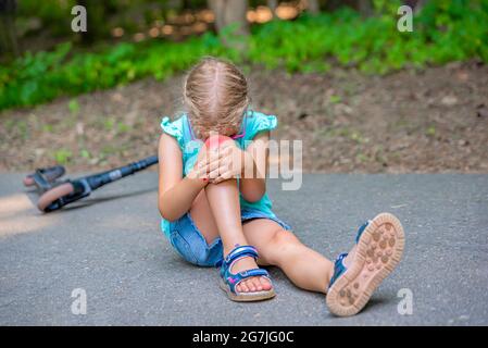 Trauma su scooter. Una bambina di cinque anni cadde su uno scooter in un parco e le ferì gravemente il ginocchio. Concetto di lesione dello scooter elettrico. Foto Stock