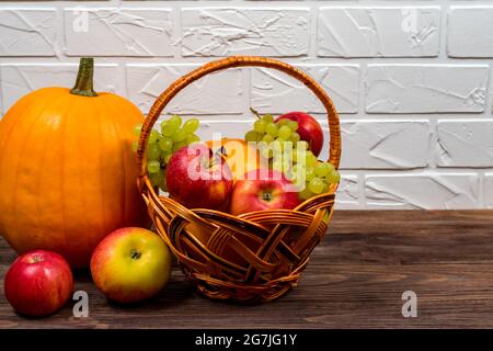 Zucche arancioni con mele e pere in un cesto di vimini su un tavolo di legno marrone sullo sfondo di un muro di mattoni bianchi. Foto Stock