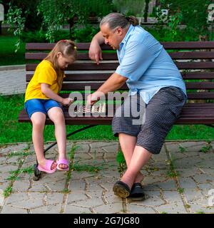 La bambina gioca a scacchi con il papà nel parco su una panchina all'aria aperta. Giorno degli Scacchi. Giochi educativi all'aperto. Sviluppo di bambini. Foto Stock