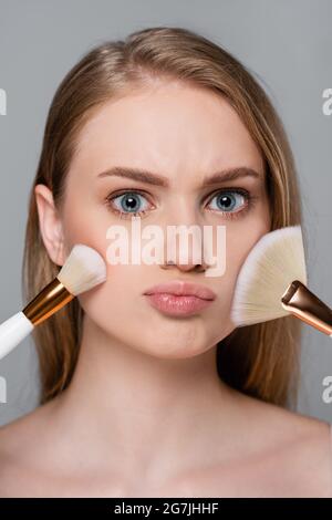 giovane donna che tiene le spazzole cosmetiche ed applica la polvere del viso isolata su grigio Foto Stock