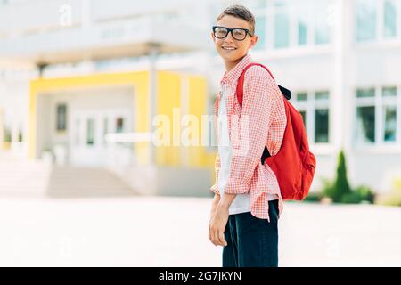 Ritorno a scuola, scolaro con occhiali con borsa, scuola elementare nel cortile, studente con libri e zaino fuori scuola Foto Stock