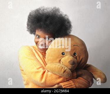 Giovane donna nera con i capelli corti che indossa un maglione giallo e che abbuffa un orsacchiotto contro un muro Foto Stock