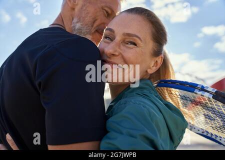 Ritratto di coppia matura attiva che guarda felice mentre si abbracciano all'aperto, pronto per l'allenamento mattutino sul campo da tennis Foto Stock