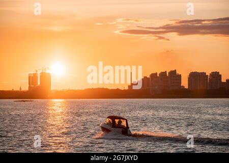 Silhouette di edifici cittadini sulla riva sullo sfondo del sole che tramonta e baglia l'acqua. Tramonto sul fiume, una barca a motore crossin Foto Stock