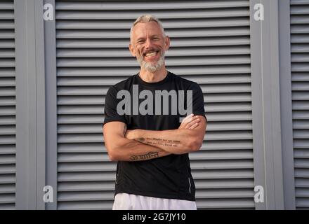 Uomo maturo di successo in perfetta forma sorridente alla telecamera mentre si alza in piedi con le braccia incrociate all'aperto su sfondo grigio Foto Stock