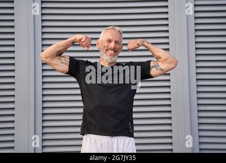 Sorridente uomo di mezza età alzò le braccia e mostra i suoi bicipiti mentre si posa all'aperto su sfondo grigio Foto Stock
