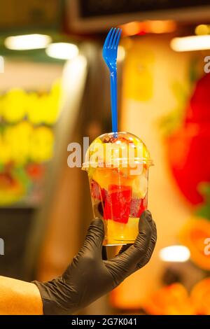 Primo piano verticale della mano di una donna con un guanto nero che tiene una tazza di plastica con frutta naturale. Foto Stock