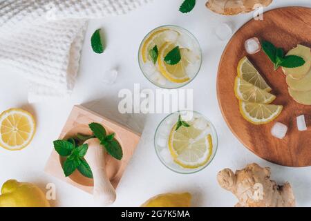 Due bicchieri con limonata e ingredienti per cucinare. Zenzero, limone, menta, ghiaccio su superficie bianca. Vassoio di legno, mortaio e pestello. Disposizione piatta Foto Stock