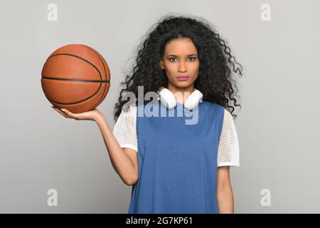 Bella giovane donna nera che bilancia un basket sulla sua mano che posa guardando la macchina fotografica con un'espressione seria e le cuffie intorno al suo collo Foto Stock