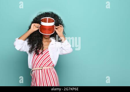 Giovane donna nera alla moda in abito a righe con un picco o visiera vintage rosso corrispondente che si affaccia su uno sfondo blu dello studio con copyspace Foto Stock