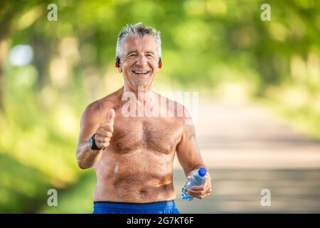 L'uomo anziano dai capelli grigi mostra i pollici mentre tiene l'acqua in bottiglia e non indossa la t-shirt dopo un allenamento nella natura. Foto Stock