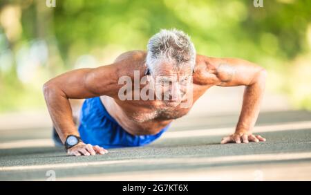 Uomo anziano che fa i pushup fuori sulla strada nella natura durante una giornata estiva soleggiata. Foto Stock