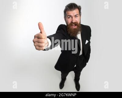 Emozionato uomo d'affari stupito in vestito nero sorridente e mostrando pollici in su. Fuga dalla crisi. Faccia divertente. Ottima idea. Ritratto di uomo eccitato gesturing Foto Stock