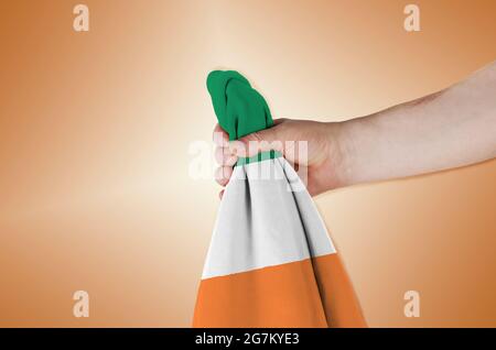 Bandiera nazionale della Repubblica d'Irlanda in mano su sfondo marrone. Uomo che detiene bandiera di un paese situato nell'Europa occidentale. Vista laterale. Foto Stock