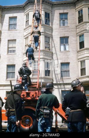 Vigili del fuoco che affrontano un blaze tetto su un edificio americano di appartamenti c.1955. Gli uomini stanno alimentando un tubo dall'apparecchio sulla scala per affrontare il problema – il fumo è visibile in alto a destra. Interessante in quei giorni jeans lavoro denim sembrava essere parte della loro uniforme. Questa immagine proviene da una vecchia trasparenza amatoriale americana del colore Kodak – una fotografia d'epoca anni '50. Foto Stock