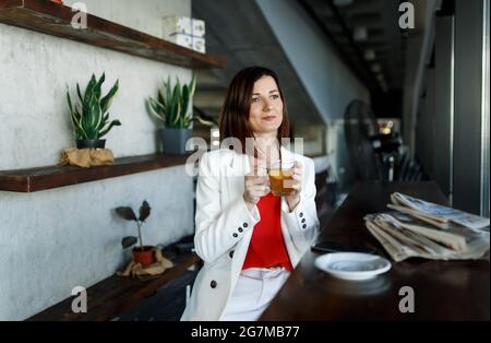 Giovane donna di bellezza millennial rilassarsi al caffè o tè freno in caffè. Indossa una giacca da ufficio bianca e una tazza con tè verde. Quotidiani sul tavolo in un moderno cafe' Place. Foto Stock