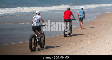 Vista posteriore di una coppia che corre in bici a ruote grasse passando un camminatore sul bordo delle acque presso la spiaggia di Fire Island New York. Foto Stock