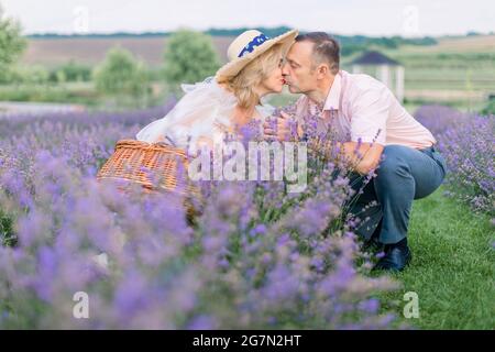 Amore e concetto di famiglia. Felice baciare la coppia matura in eleganti abiti eleganti, seduti in un bellissimo campo di lavanda fiorito e godendosi il momento Foto Stock