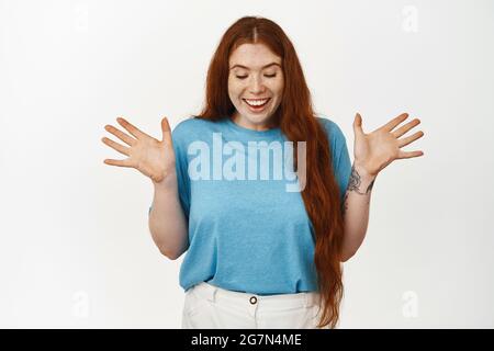 Sorpresa. Felice ragazza rossa festeggiando, scuotendo le mani e guardando giù con espressione allegra ed eccitata, vincendo qualcosa, regalo aperto, in piedi Foto Stock