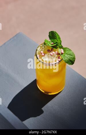 Bella limonata rinfrescante decorata con caramelle rotonde rosa e menta. Cocktail giallo con ghiaccio sul balcone. Drink estivo. Foto verticale. Foto Stock