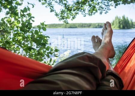 L'uomo a piedi nudi si rilassa disteso in un'amaca all'ombra degli alberi, con una magnifica vista sul lago, in una giornata estiva. Foto Stock