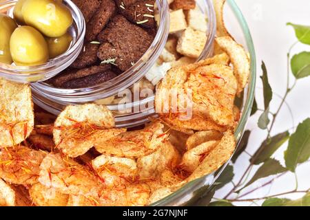 Selezione focalizzata, composizione alimentare con spuntini salati: Cracker di segala, cracker di grano, patatine con spezie e olive verdi in ciotole di vetro su un bianco Foto Stock